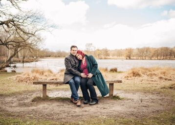Expectant couple sat on a bench in front of a lake in Richmond Park