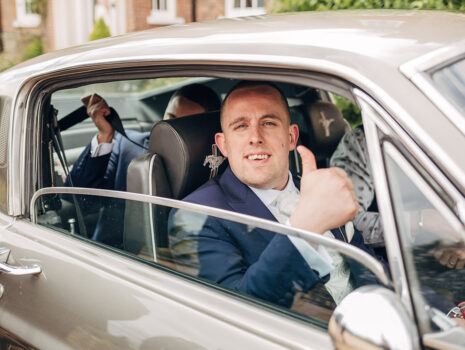 A groom giving the thumbs up as he drives to the wedding ceremony in a vintage Ford Mustang