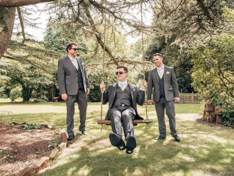A groom and two groomsmen having fun on a swing in the garden of the groom's parents
