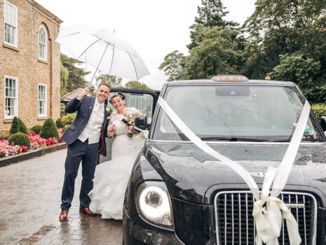 A wedding couple posing with an umbrella next to their black car wedding car