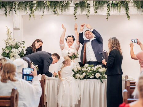 A wedding couple cheering and clapping while witnesses sign the marriage register during their ceremony