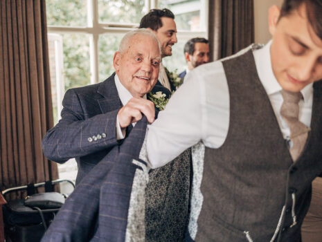 The bride's grandfather helping a groom put his jacket on while getting ready